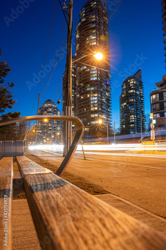 Canvas Print Selective focus on a bus stop bench, with light trails streak down a busy city s