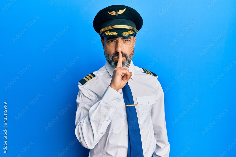 Middle age man with beard and grey hair wearing airplane pilot uniform asking to be quiet with finger on lips. silence and secret concept. Middle age man with beard and grey hair wearing airplane pilot uniform asking to be quiet with finger on lips. silence and secret concept.