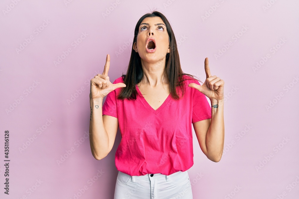 Young hispanic woman wearing casual clothes amazed and surprised looking up and pointing with fingers and raised arms.