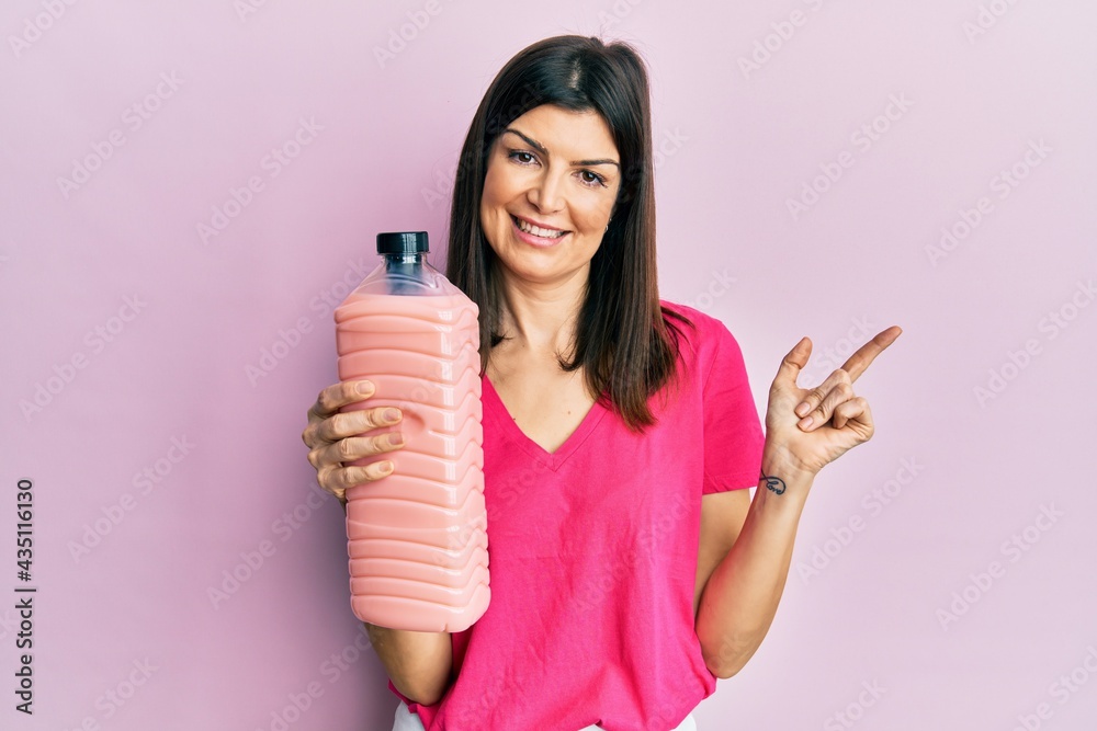Young hispanic woman holding laundry softener bottle smiling happy pointing with hand and finger to the side Young hispanic woman holding laundry softener bottle smiling happy pointing with hand and finger to the side