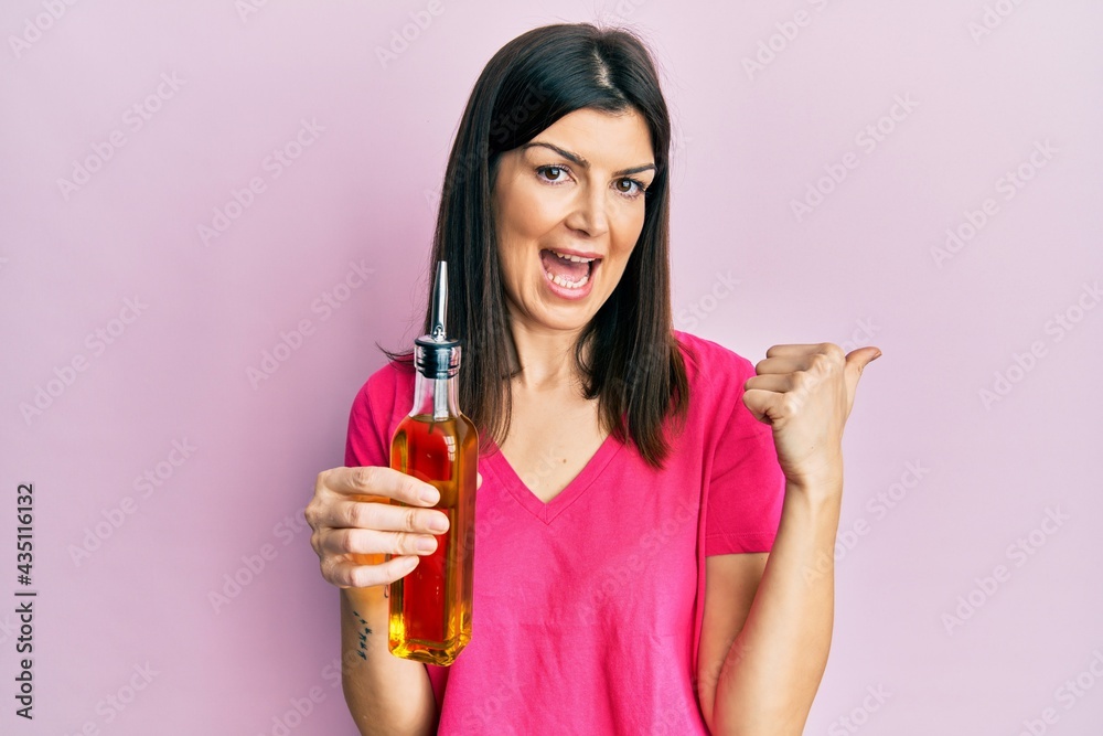 Young hispanic woman holding olive oil can pointing thumb up to the side smiling happy with open mouth Young hispanic woman holding olive oil can pointing thumb up to the side smiling happy with open mouth