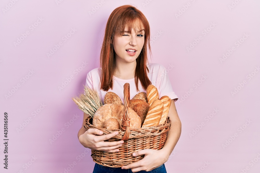 Redhead young woman holding wicker basket with bread winking looking at the camera with sexy expression, cheerful and happy face.