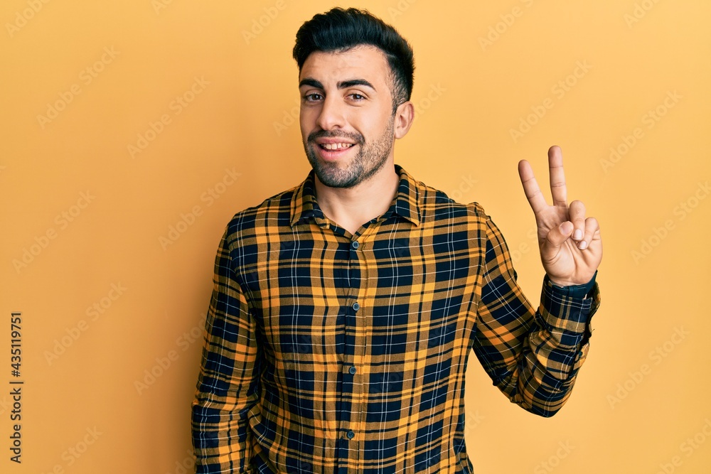 Naklejka premium Young hispanic man wearing casual clothes smiling looking to the camera showing fingers doing victory sign. number two.