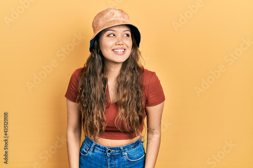Young hispanic girl wearing casual clothes and hat looking away to side with smile on face, natural expression. laughing confident.