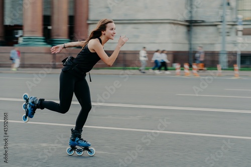 Holidays and active lifestyle concept. Slim healthy European woman rollerskates on high speed photogaphed in motion dressed in black clothing poses against blurred street background on road.