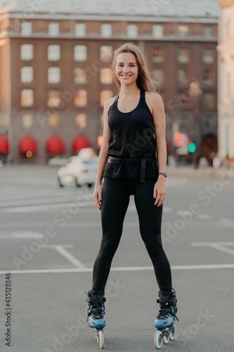 Positive young female model in sportsclothes rides on blades enjoys leisure activities poses at urban place against blurred background stands in full length. Active lifestyle and rollerblading concept © VK Studio