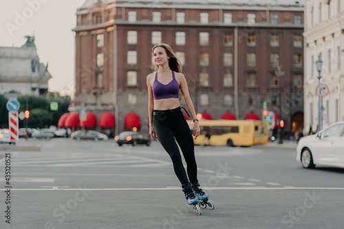 Full length shot of sporty slim woman in active wear rides on blades enjoys outdoor fitness activity during warm summer day poses at urban place on asphalt. Rollerblading and leisure concept