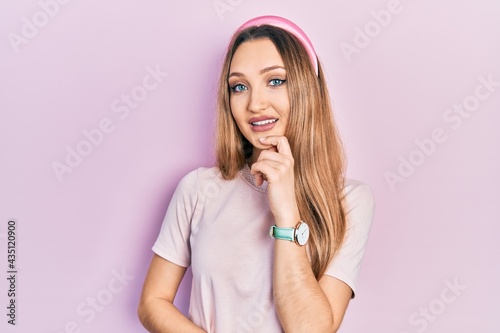 Young blonde girl wearing casual clothes looking confident at the camera smiling with crossed arms and hand raised on chin. thinking positive.