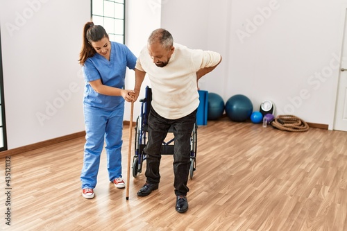 Senior man sitting on wheelchair being assited by geriatric nurse, handicapped mature man getting help to stand up photo