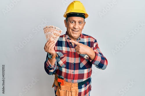 Senior hispanic man wearing handyman uniform holding turkish liras smiling happy pointing with hand and finger