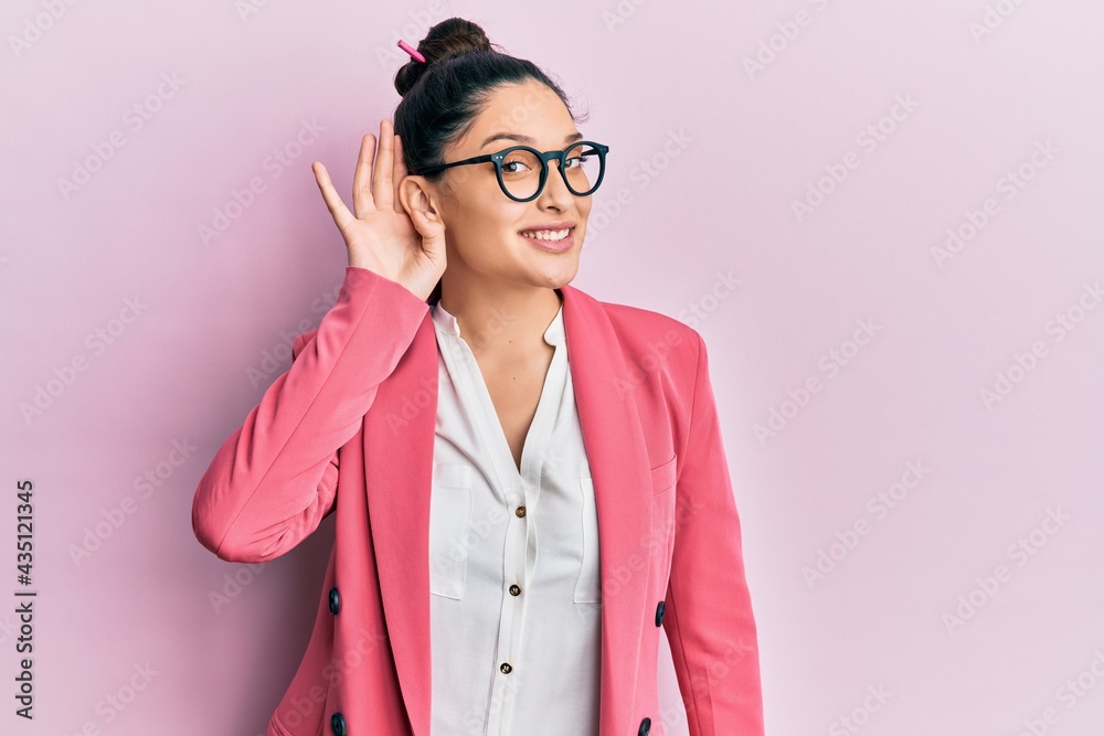 Beautiful middle eastern woman wearing business jacket and glasses smiling with hand over ear listening an hearing to rumor or gossip. deafness concept.