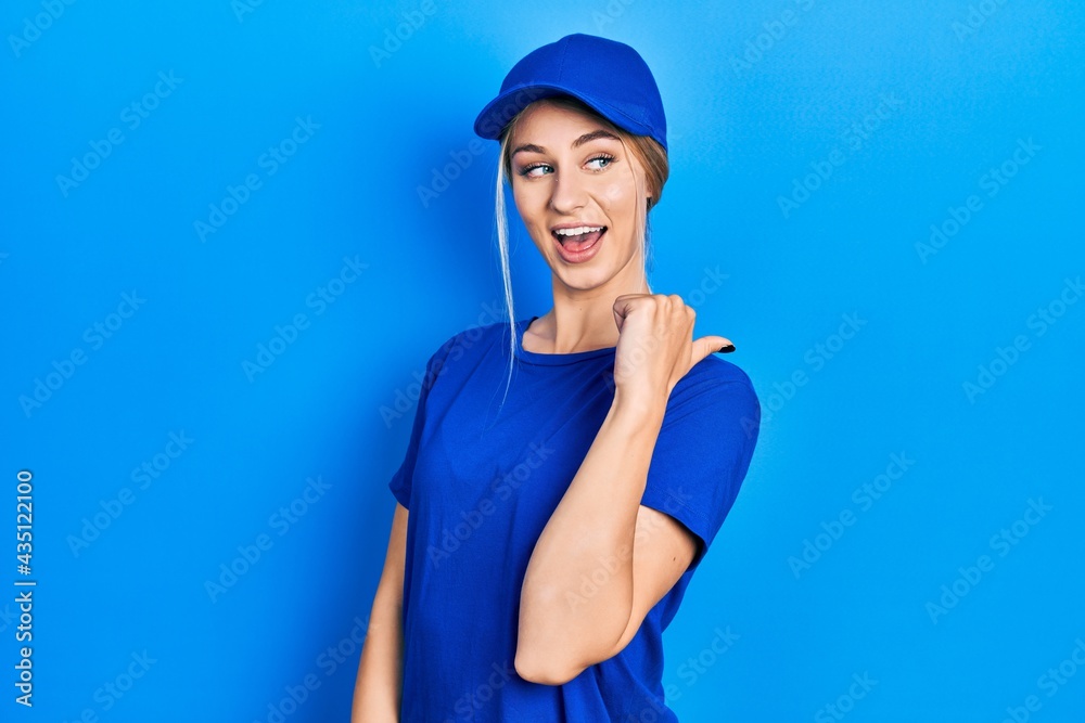 Young caucasian woman wearing courier uniform wearing cap smiling with happy face looking and pointing to the side with thumb up. Young caucasian woman wearing courier uniform wearing cap smiling with happy face looking and pointing to the side with thumb up.