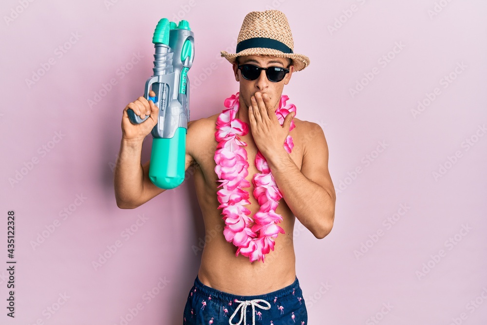 Young caucasian man wearing swimsuit and hawaiian lei holding watergun covering mouth with hand, shocked and afraid for mistake. surprised expression Young caucasian man wearing swimsuit and hawaiian lei holding watergun covering mouth with hand, shocked and afraid for mistake. surprised expression