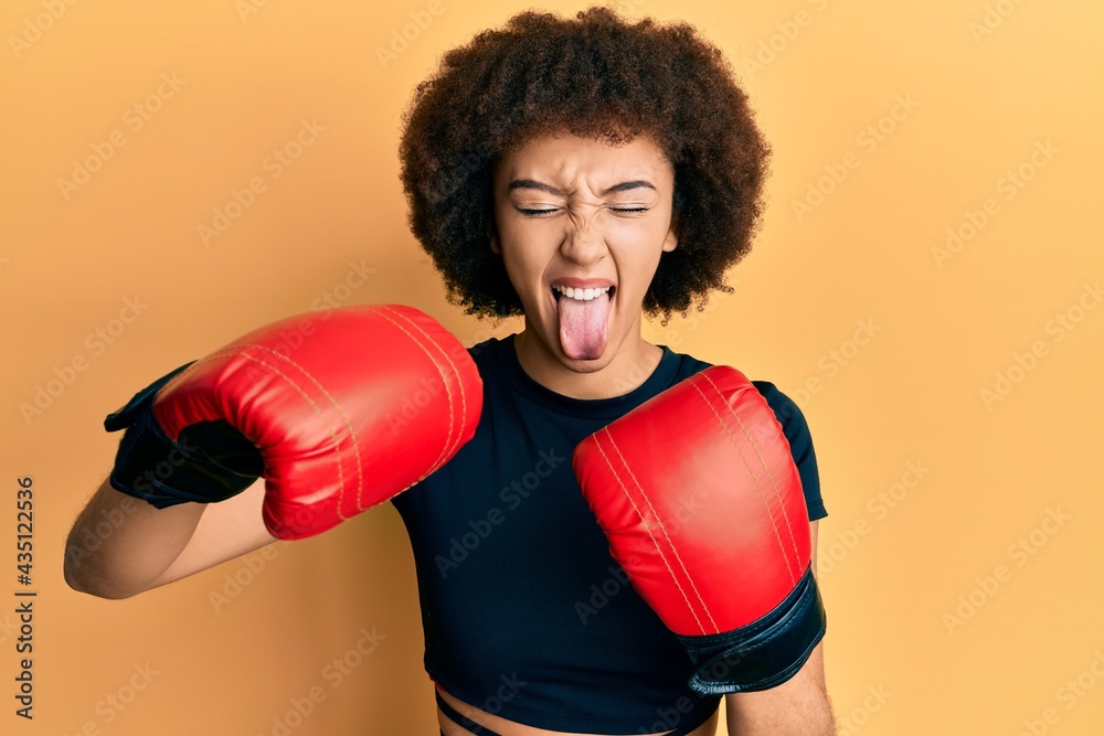 Young hispanic sporty girl using boxing gloves sticking tongue out happy with funny expression.