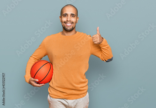 Hispanic adult man holding basketball ball smiling happy and positive, thumb up doing excellent and approval sign