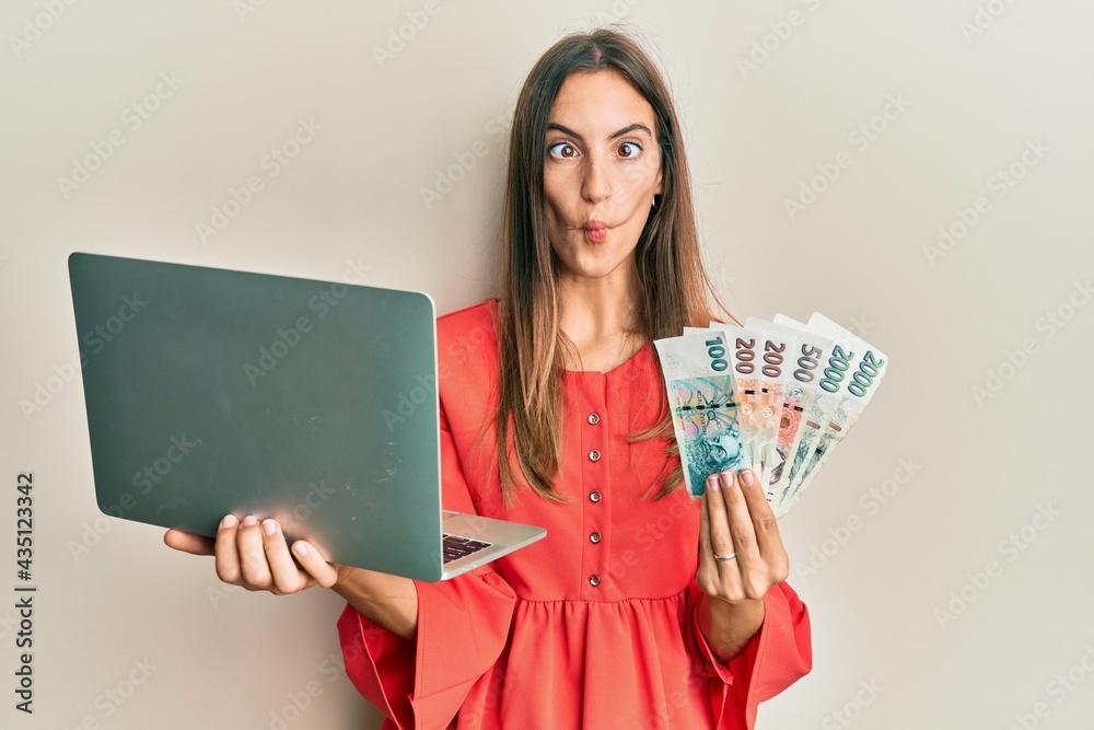 Young beautiful woman holding laptop and czech koruna banknotes making fish face with mouth and squinting eyes, crazy and comical. Young beautiful woman holding laptop and czech koruna banknotes making fish face with mouth and squinting eyes, crazy and comical.
