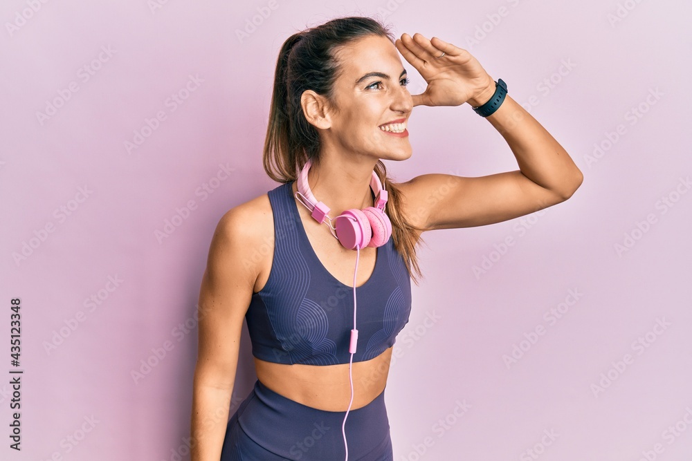 Young beautiful woman wearing gym clothes and using headphones very happy and smiling looking far away with hand over head. searching concept.