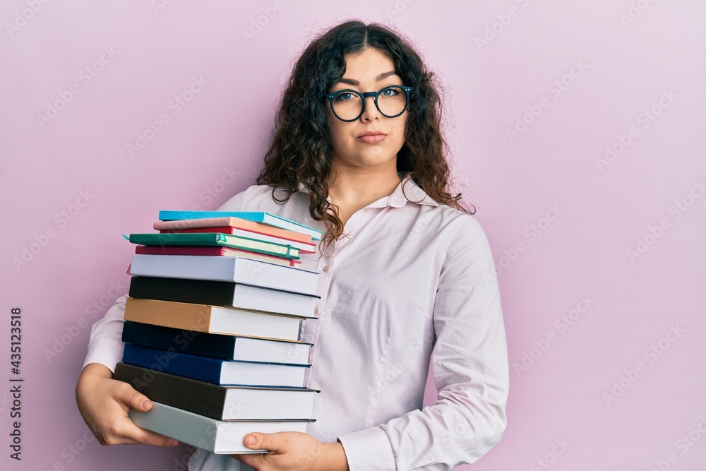 Young brunette woman with curly hair holding a pile of books relaxed with serious expression on face. simple and natural looking at the camera. Young brunette woman with curly hair holding a pile of books relaxed with serious expression on face. simple and natural looking at the camera.