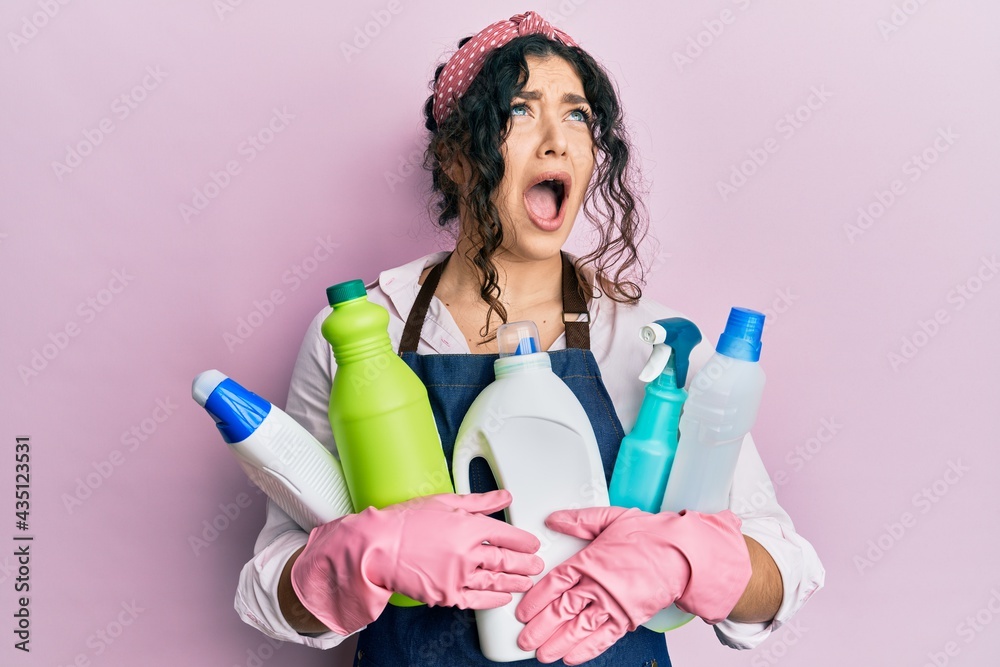 Young brunette woman with curly hair wearing cleaner apron holding cleaning products angry and mad screaming frustrated and furious, shouting with anger looking up. Young brunette woman with curly hair wearing cleaner apron holding cleaning products angry and mad screaming frustrated and furious, shouting with anger looking up.