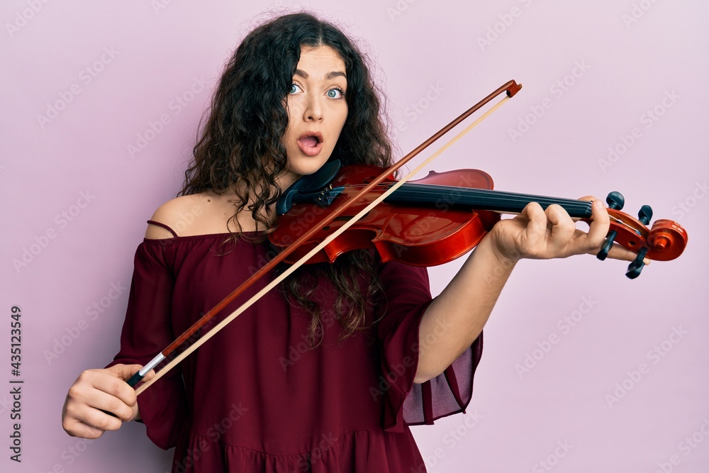Young brunette musician woman with curly hair playing violin afraid and shocked with surprise and amazed expression, fear and excited face. Young brunette musician woman with curly hair playing violin afraid and shocked with surprise and amazed expression, fear and excited face.