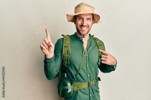 Handsome man with beard wearing explorer hat and backpack with a big smile on face, pointing with hand finger to the side looking at the camera.