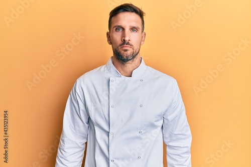 Handsome man with beard wearing professional cook uniform relaxed with serious expression on face. simple and natural looking at the camera.