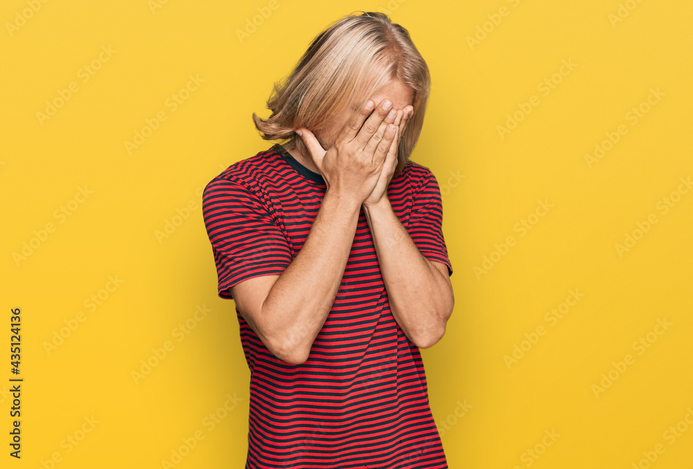 Caucasian man with blond long hair wearing casual striped t shirt with sad expression covering face with hands while crying. depression concept. Caucasian man with blond long hair wearing casual striped t shirt with sad expression covering face with hands while crying. depression concept.