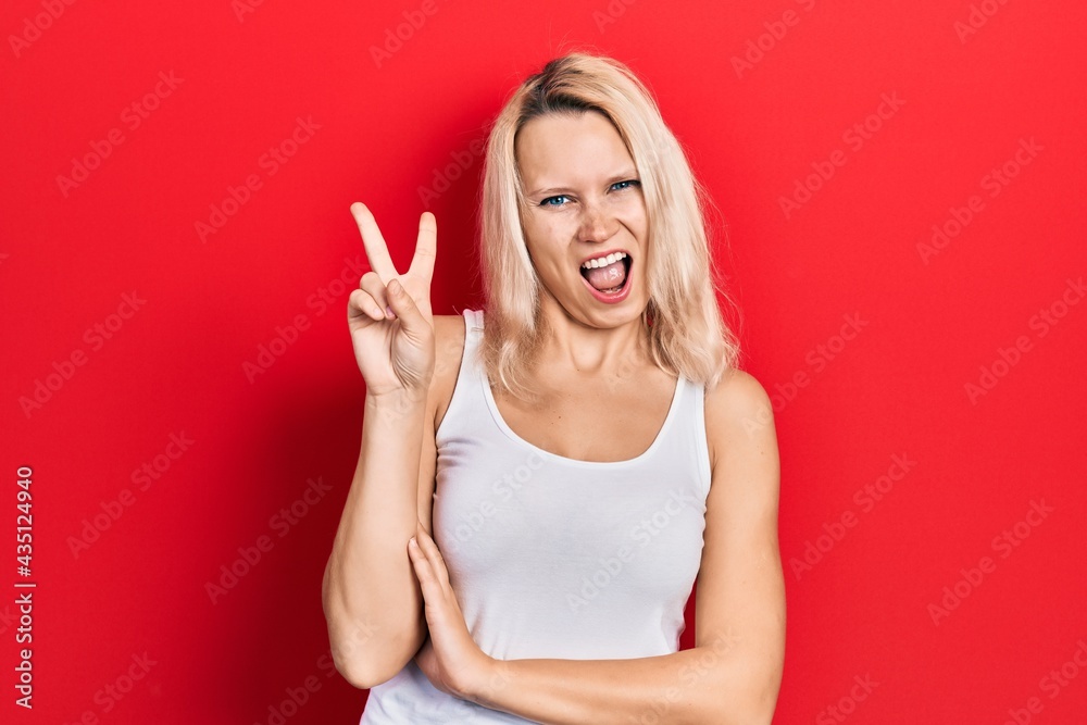 Beautiful caucasian blonde woman wearing casual white t shirt smiling with happy face winking at the camera doing victory sign. number two. Beautiful caucasian blonde woman wearing casual white t shirt smiling with happy face winking at the camera doing victory sign. number two.