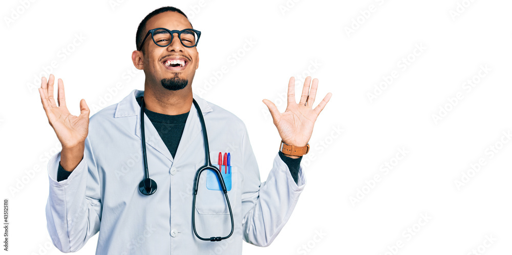 Young african american man wearing doctor uniform and stethoscope celebrating crazy and amazed for success with arms raised and open eyes screaming excited. winner concept Young african american man wearing doctor uniform and stethoscope celebrating crazy and amazed for success with arms raised and open eyes screaming excited. winner concept