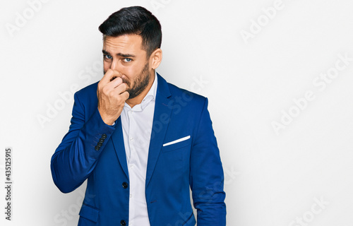 Young hispanic man wearing business jacket smelling something stinky and disgusting, intolerable smell, holding breath with fingers on nose. bad smell