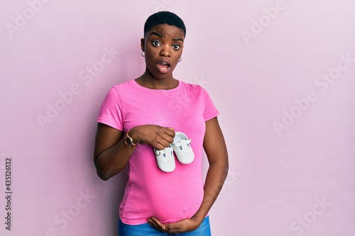 Young african american woman expecting a baby holding shoes in shock face, looking skeptical and sarcastic, surprised with open mouth