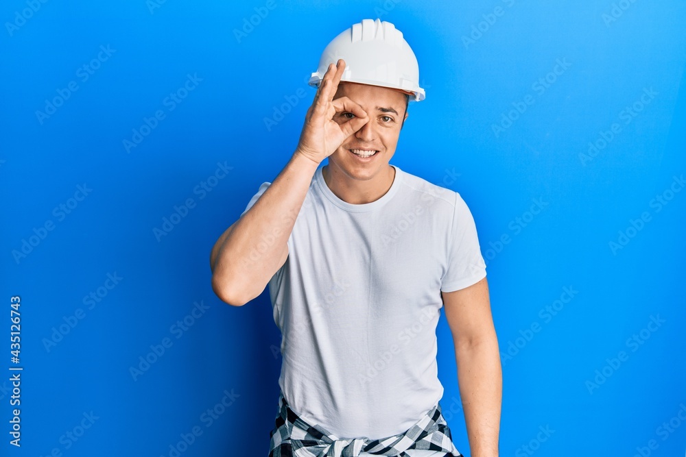 Handsome young man wearing builder uniform and hardhat doing ok gesture with hand smiling, eye looking through fingers with happy face.