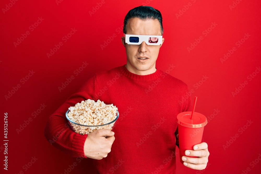 Handsome young man eating popcorn and soda wearing 3d glasses in shock face, looking skeptical and sarcastic, surprised with open mouth