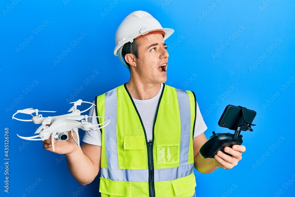 Handsome young man wearing safety hard using drone and control angry and mad screaming frustrated and furious, shouting with anger looking up. Handsome young man wearing safety hard using drone and control angry and mad screaming frustrated and furious, shouting with anger looking up.
