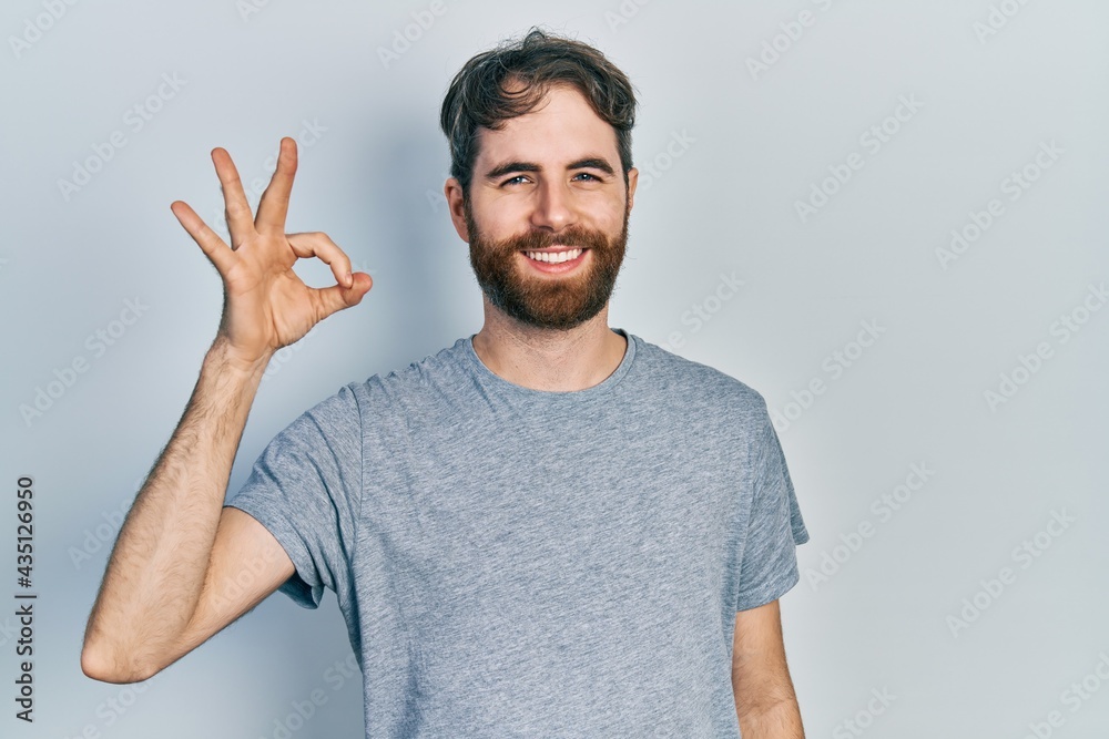 Caucasian man with beard wearing casual grey t shirt smiling positive doing ok sign with hand and fingers. successful expression. Caucasian man with beard wearing casual grey t shirt smiling positive doing ok sign with hand and fingers. successful expression.