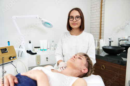 Attractive young doctor dermatologist sitting near a spa center client on the background of professional medical equipments in contemporary beauty SPA clinic