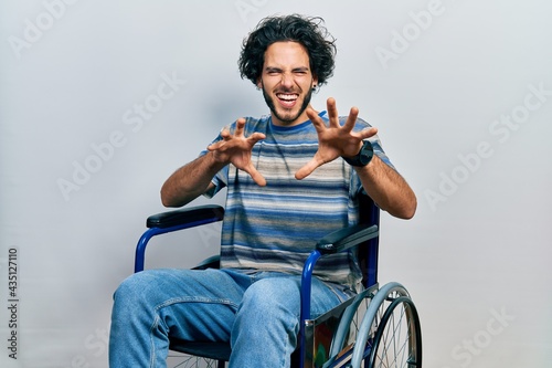 Handsome hispanic man sitting on wheelchair smiling funny doing claw gesture as cat, aggressive and sexy expression