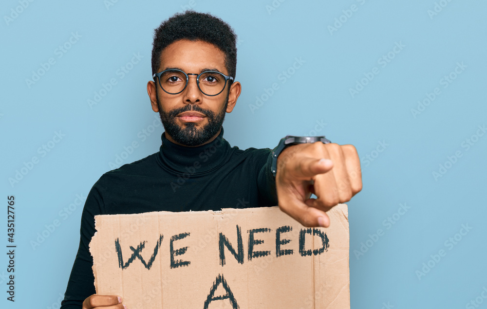 Young african american man holding we need a change banner pointing with finger to the camera and to you, confident gesture looking serious Young african american man holding we need a change banner pointing with finger to the camera and to you, confident gesture looking serious