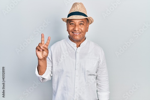 Middle age latin man wearing casual clothes and hat smiling with happy face winking at the camera doing victory sign. number two.
