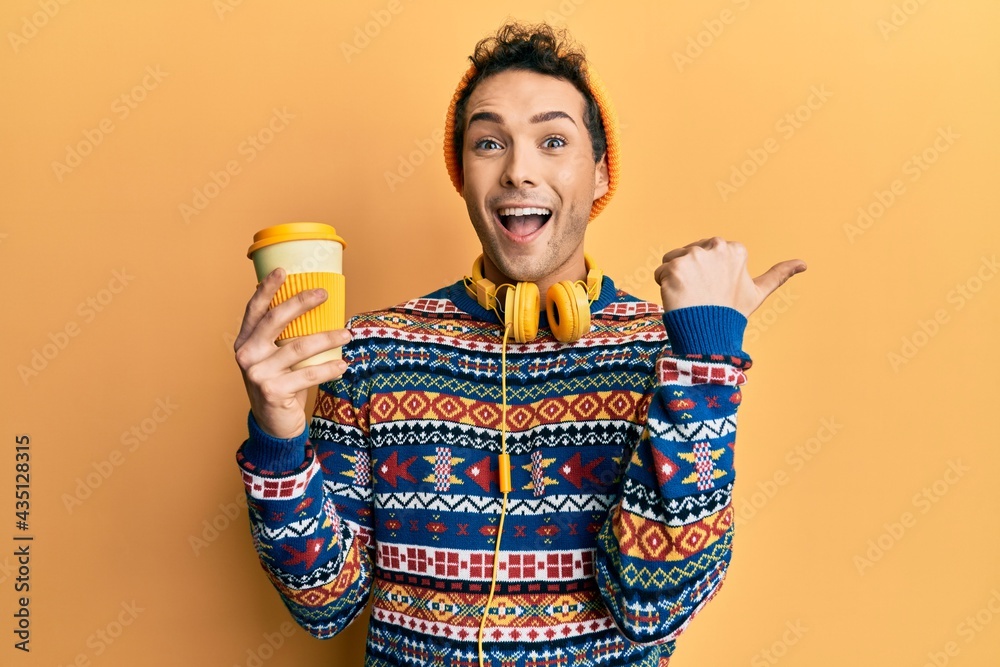 Young handsome man drinking cup of coffee wearing headphones pointing thumb up to the side smiling happy with open mouth Young handsome man drinking cup of coffee wearing headphones pointing thumb up to the side smiling happy with open mouth