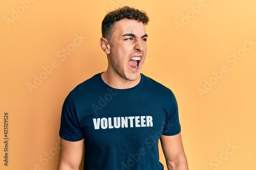 Hispanic young man wearing volunteer t shirt angry and mad screaming frustrated and furious, shouting with anger. rage and aggressive concept.