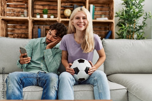 Woman smiling happy watching soccer match and boyfriend boring using smartphone at home.