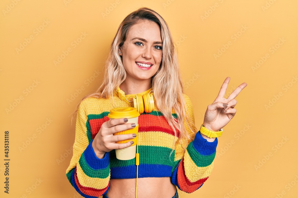 Beautiful young blonde woman drinking cup of coffee wearing headphones smiling looking to the camera showing fingers doing victory sign. number two. Beautiful young blonde woman drinking cup of coffee wearing headphones smiling looking to the camera showing fingers doing victory sign. number two.