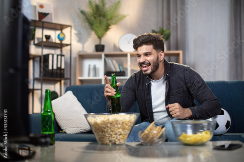 Excited indian man with chips and beer sitting on cozy sofa and watching sport TV. Young bearded guy supporting favorite football team during world cup. © sofiko14