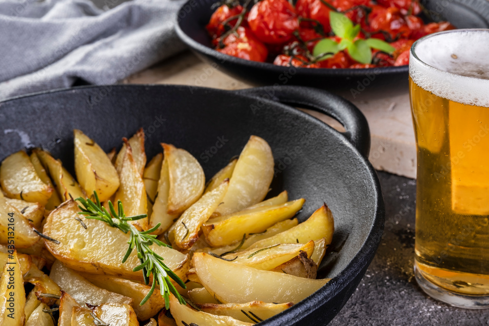 Roasted potatoes with rosemary in iron casserole and plate of confit tomatoes on dark background