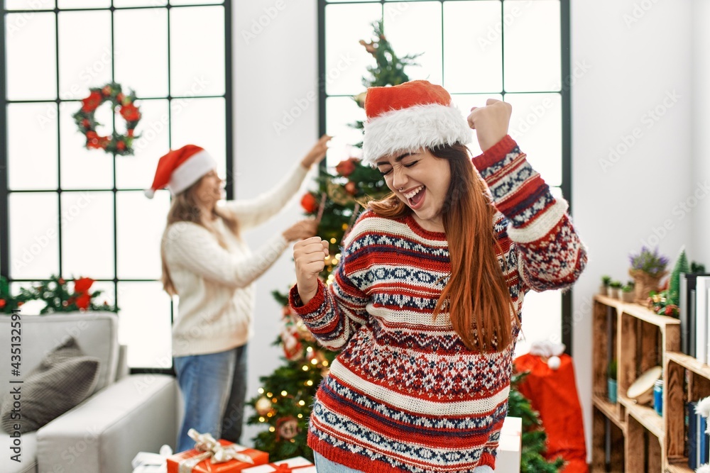 Beautiful couple at home standing by christmas tree very happy and excited doing winner gesture with arms raised, smiling and screaming for success. celebration concept.