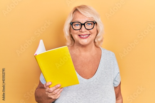 Middle age blonde woman reading a book looking positive and happy standing and smiling with a confident smile showing teeth