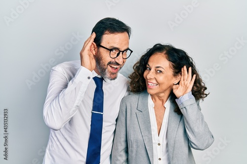 Middle age couple of hispanic woman and man wearing business office uniform smiling with hand over ear listening an hearing to rumor or gossip. deafness concept.