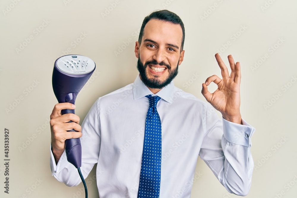 Young business man with beard holding electric steam iron doing ok sign with fingers, smiling friendly gesturing excellent symbol Young business man with beard holding electric steam iron doing ok sign with fingers, smiling friendly gesturing excellent symbol