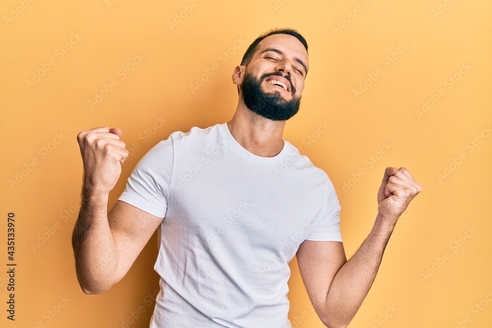 Young man with beard wearing casual white t shirt very happy and excited doing winner gesture with arms raised, smiling and screaming for success. celebration concept. Young man with beard wearing casual white t shirt very happy and excited doing winner gesture with arms raised, smiling and screaming for success. celebration concept.